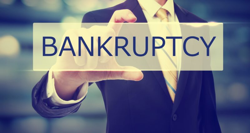 A man in a black suit holding up a rectangular sign that reads bankruptcy with one hand