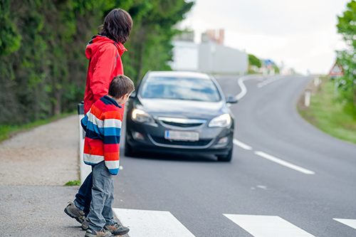 A parent and child cautiously walk toward a crosswalk as a grey sedan drives toward them.