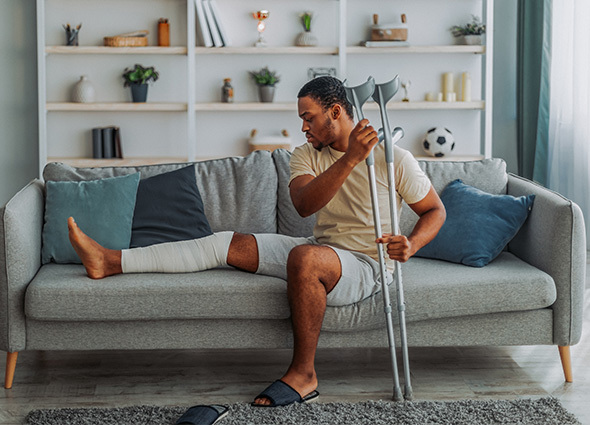 A man with his leg wrapped and a pair of crutches sits on a couch in his home